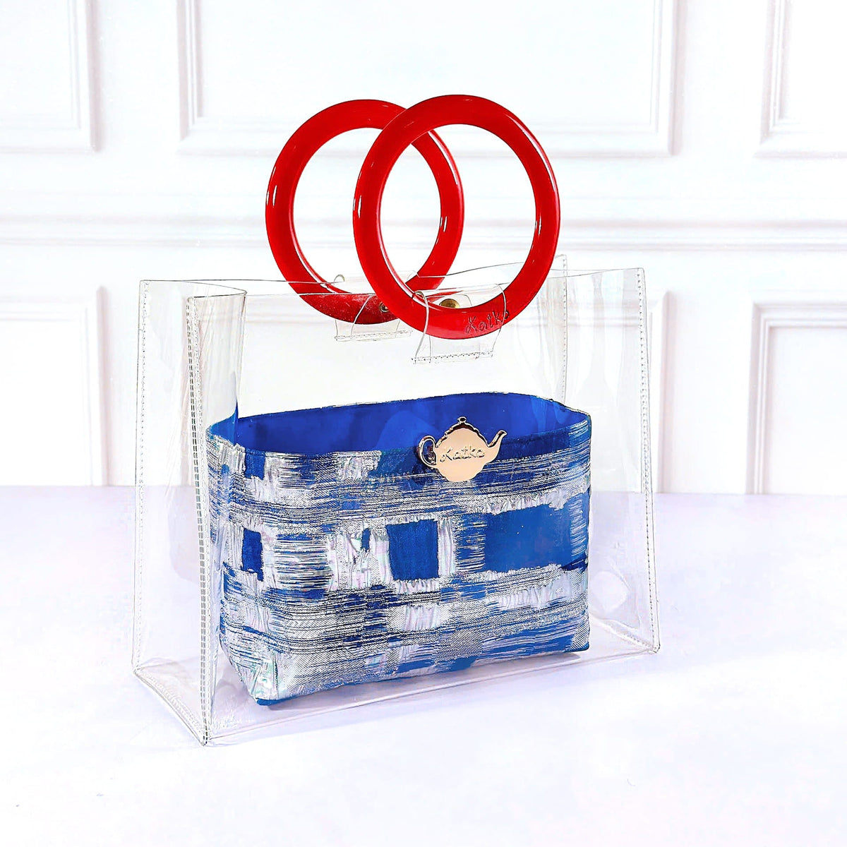 A clear acrylic handbag with a blue and white patterned fabric pouch, featuring two large red interlocking ring handles, set against a white paneled background.