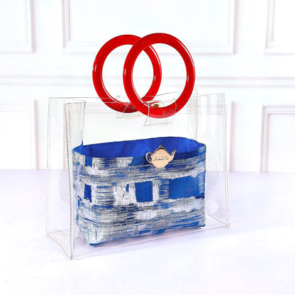 A clear acrylic handbag with a blue and white patterned fabric pouch, featuring two large red interlocking ring handles, set against a white paneled background.