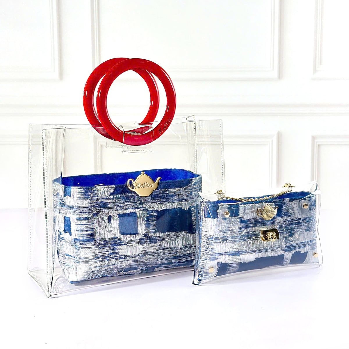 Two clear plastic handbags with blue and white patterned fabric interiors, displayed against a white paneled wall. One larger bag features a prominent red handle, and both bags have gold-tone hardware.