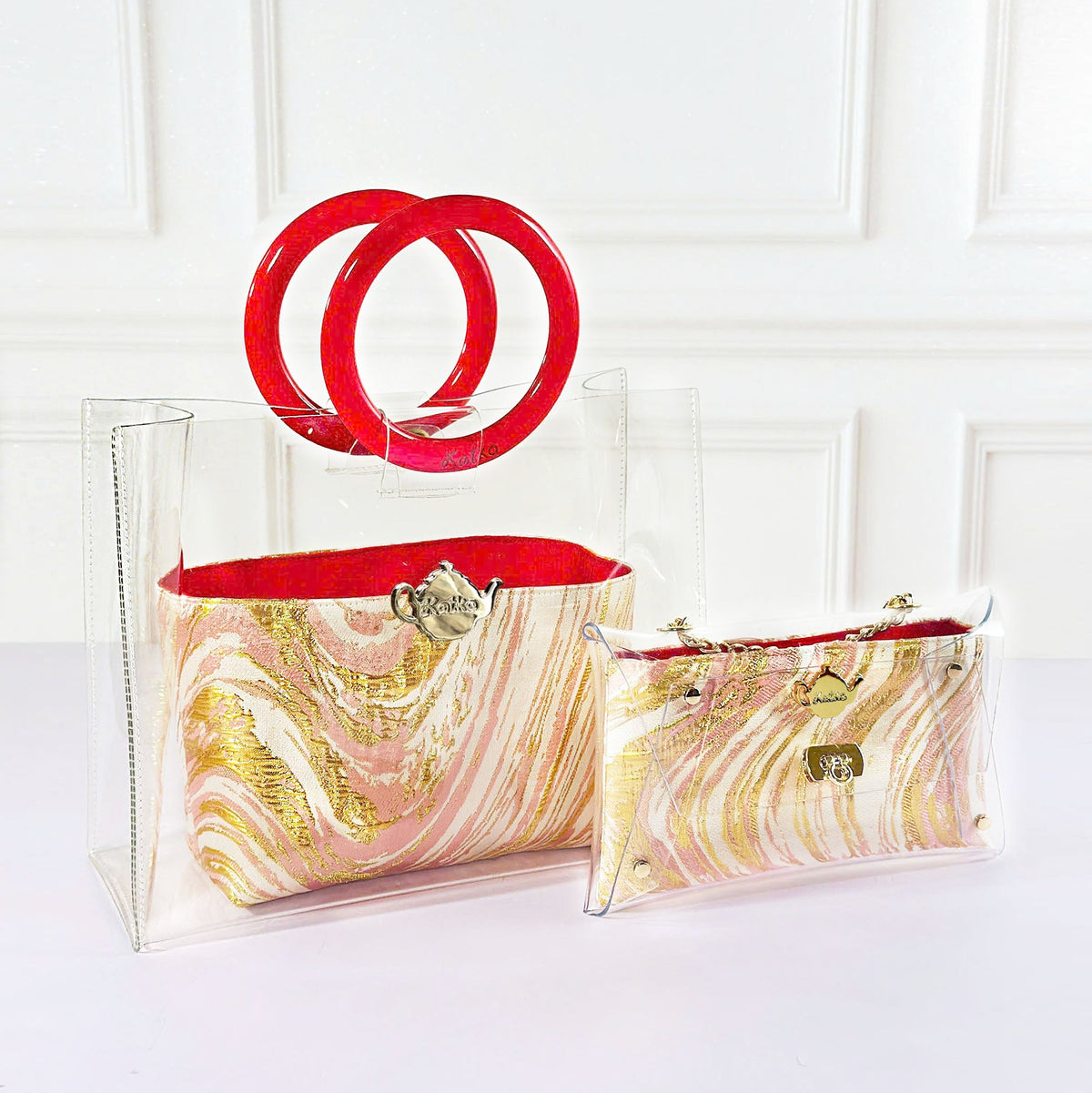 Two clear acrylic handbags with a pink, gold, and white marble pattern, one featuring a large red double-ring handle and the other a red zipper closure, displayed against a white paneled wall.