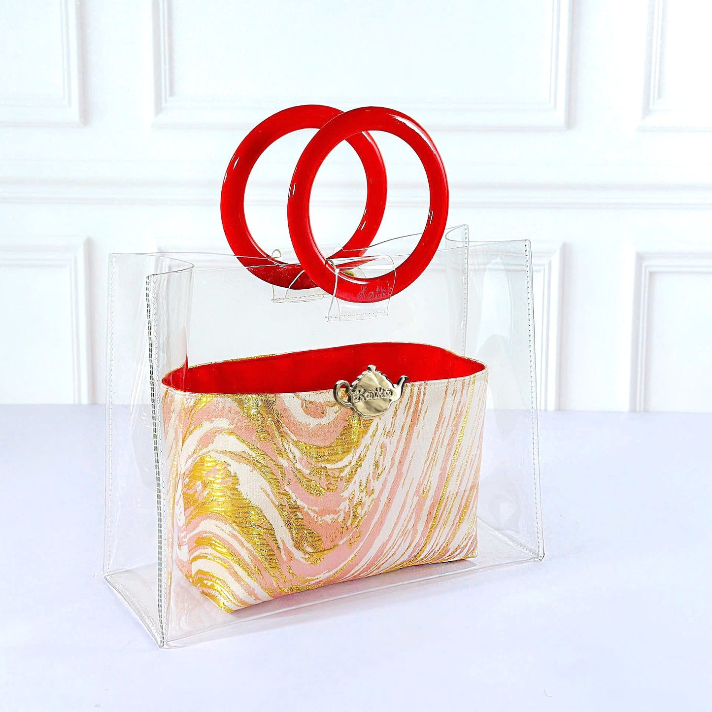 A clear acrylic tote bag with a pink, gold, and white marbled patterned pouch inside, featuring a bright red interior lining and two interlocking red handles, set against a white paneled wall.