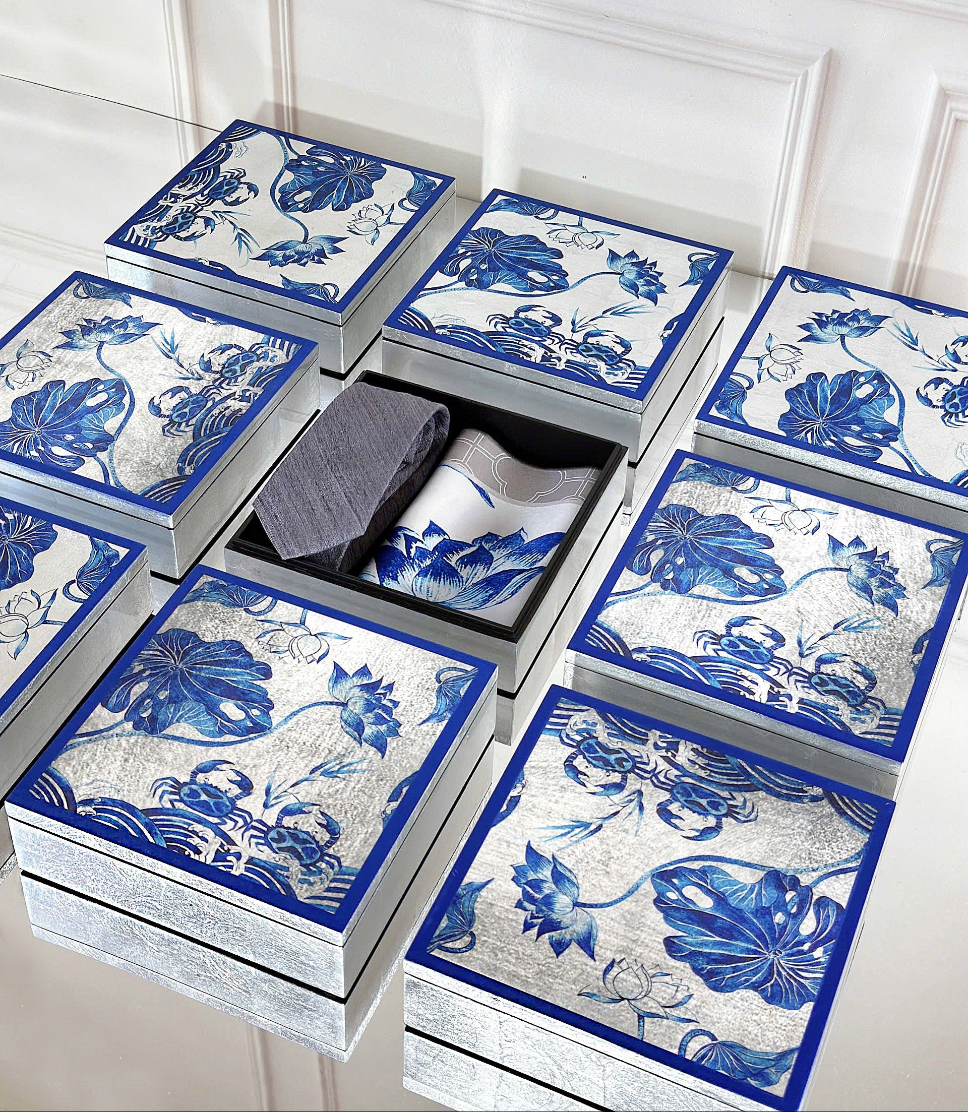 A set of six silver drawers with blue and white floral porcelain-style patterns, arranged on a white paneled surface with one drawer open to reveal folded gray and white garments inside.